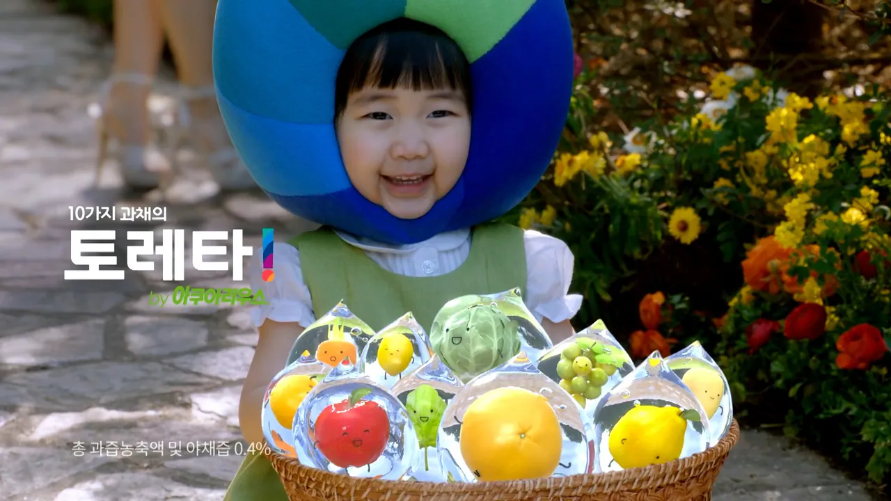 Child wearing a blue-and-green globe-shaped headpiece, smiling and holding a basket filled with CG fruit-and-vegetable droplet characters, in a flower garden