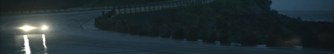 Wet night road along a rocky coastline, with a car’s headlights closer and reflecting off the road