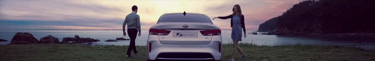 White Kia K5 Plug-in Hybrid seen from behind at a coastal overlook at dusk, with two people walking toward it and a pink-and-blue sunset over the sea