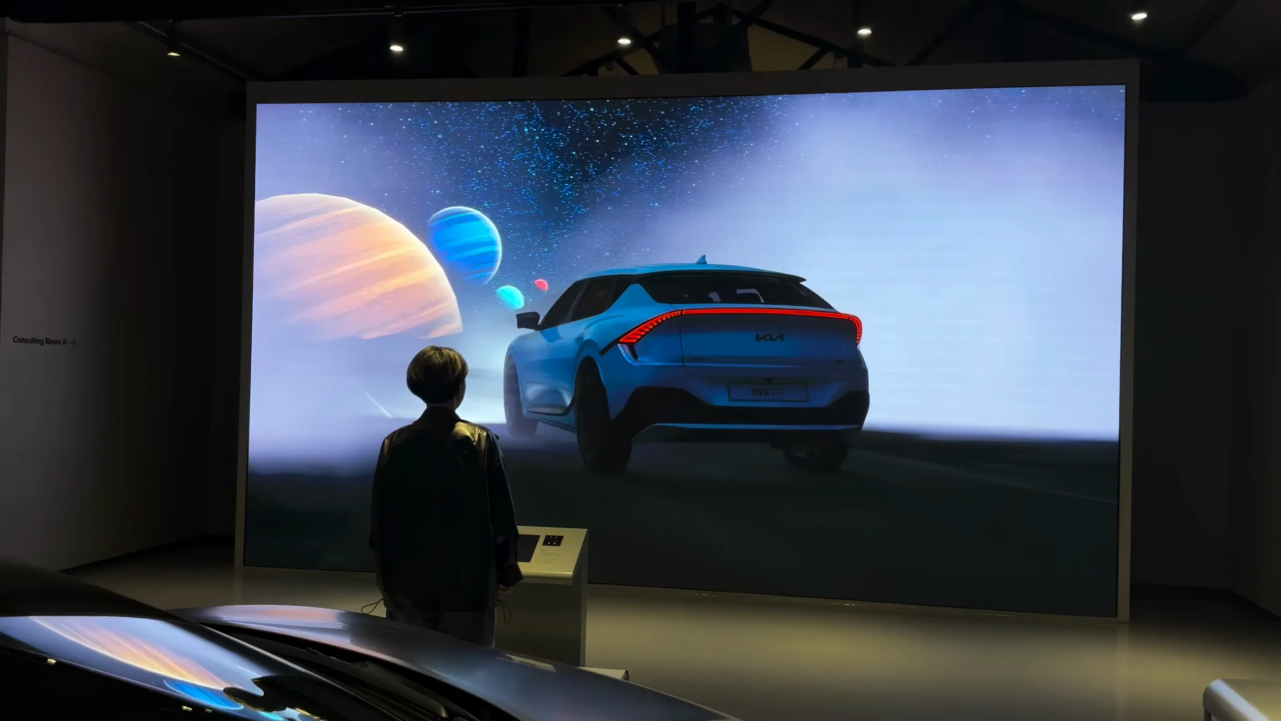 Visitor at a kiosk in front of a large LED wall showing a Kia electric car driving toward planets and a starfield